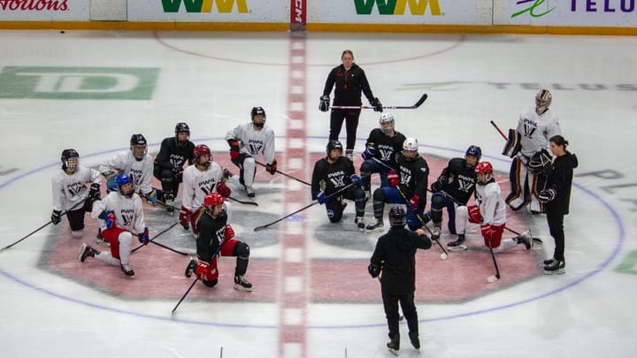 Hockey players in PWHL jerseys take a knee at centre ice infront of coach Carla MacLeod.