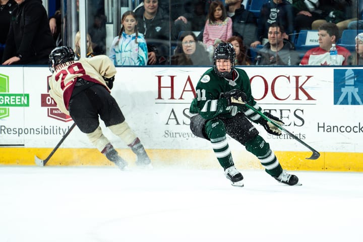 Alina Müller, wearing a green home uniform, and Marie-Philip Poulin, wearing a cream away uniform, turn towards a play.