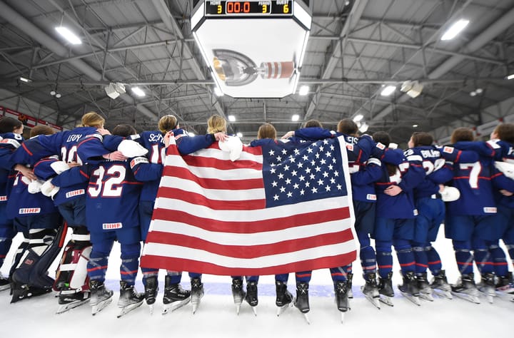 Team USA stands arm-in-arm, on the blue line. The American flag hangs over some backs.