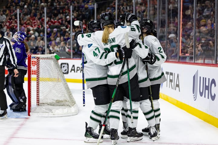 Boston celebrates a goal against Minnesota. They are all in a tight group hug and wearing white away uniforms.
