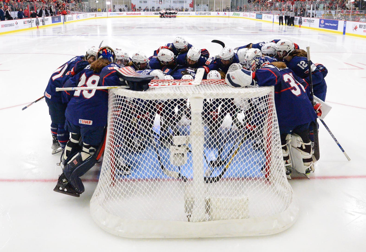 Team USA huddles around the net before the 2023 IIHF World Championship gold medal game. They are in navy blue uniforms.