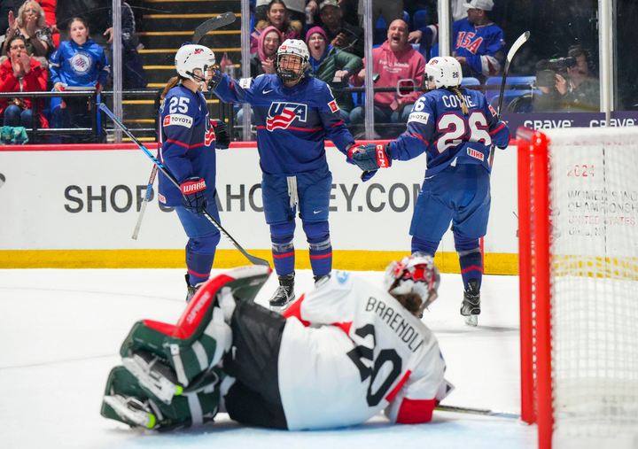 Winn is about to be in a group hug with two teammates, all wearing blue. Brändli (in white) lays on the ice in front of them.