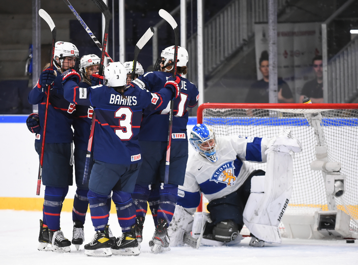 Team USA players celebrate a goal against Finland with a group hug. The Finnish netminder is kneeling in the background.