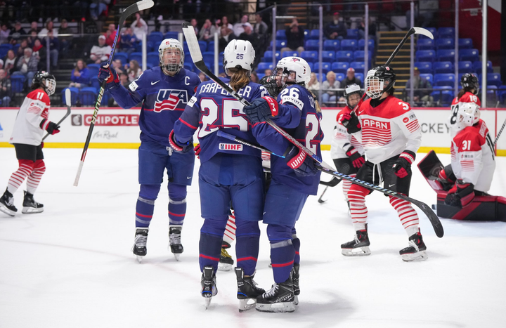 Carpenter celebrates her goal with a group hug featuring Coyne Schofield and Knight. They are wearing blue uniforms.