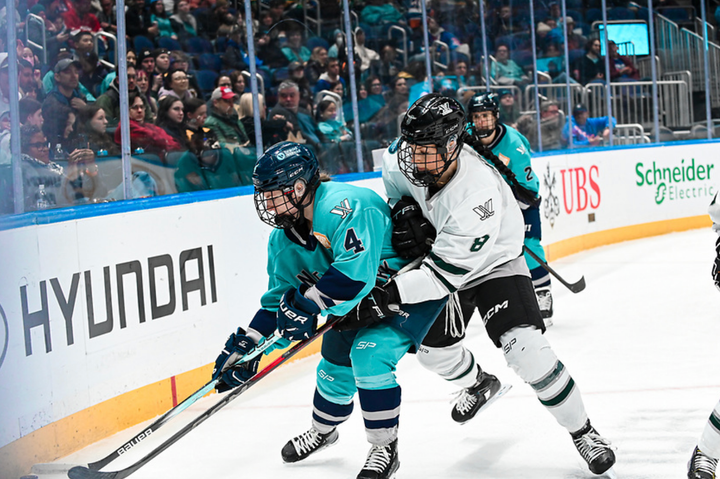 Adzija (right in white) battles for the puck with Baker (left in teal) in the corner. Adzija is reaching around Baker.