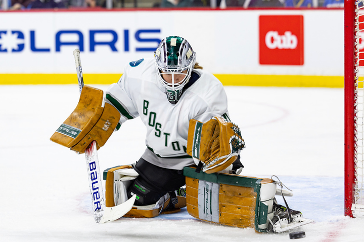 Frankel kicks the puck to her left from the butterfly position. She is wearing a white uniform and light brown pads.