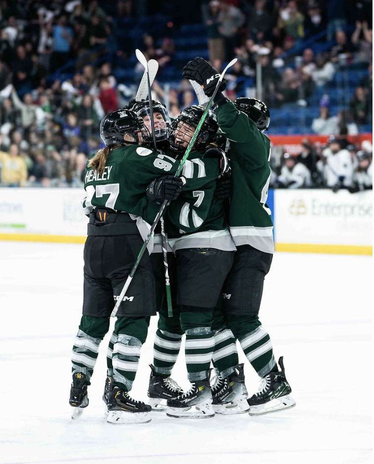 Five Boston players celebrate with a hug at the top of a face-off circle. They're smiling and wearing green home uniforms.