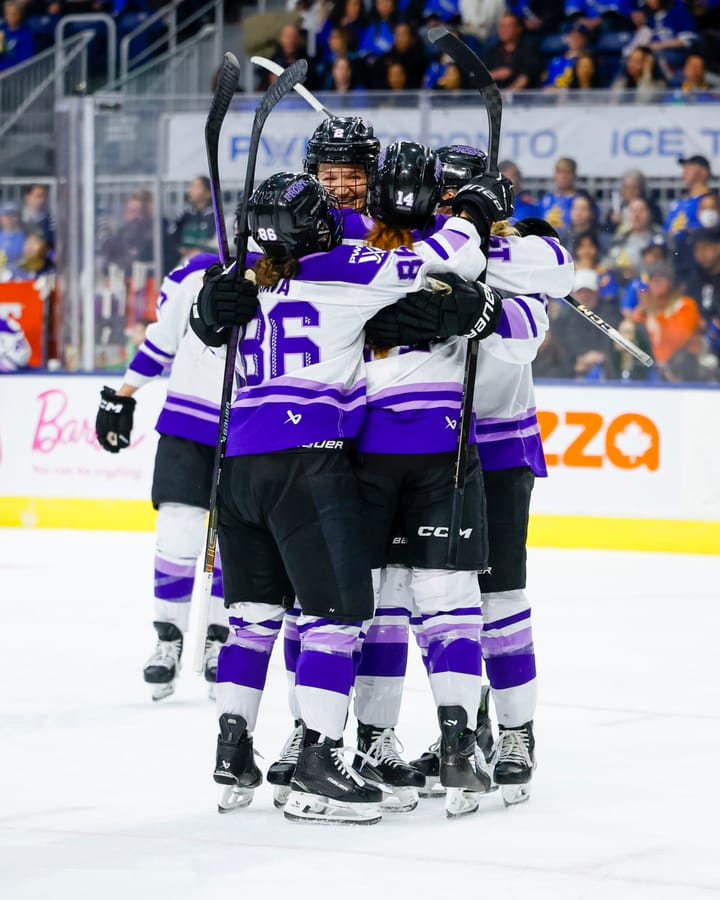 Minnesota players celebrate with a tight group hug by the faceoff circle. They are all wearing white away uniforms.