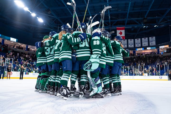 The Boston Fleet celebrate a win with a tight group hug at center ice. They are raising their sticks and wearing green home uniforms.