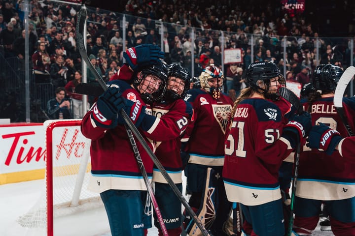 Multiple Montréal Victoire players in red jerseys gather around the net smiling and congratulating each other
