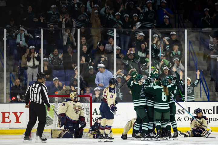 Five members of the Fleet celebrate a goal with a tight group hug to the left of Ann-Renée Desbiens. The Fleet are in green home uniforms, while the Victoire are in cream away ones.
