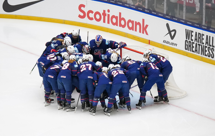 Team USA forms a tight huddle around their crease before the game. They are all wearing blue home uniforms.